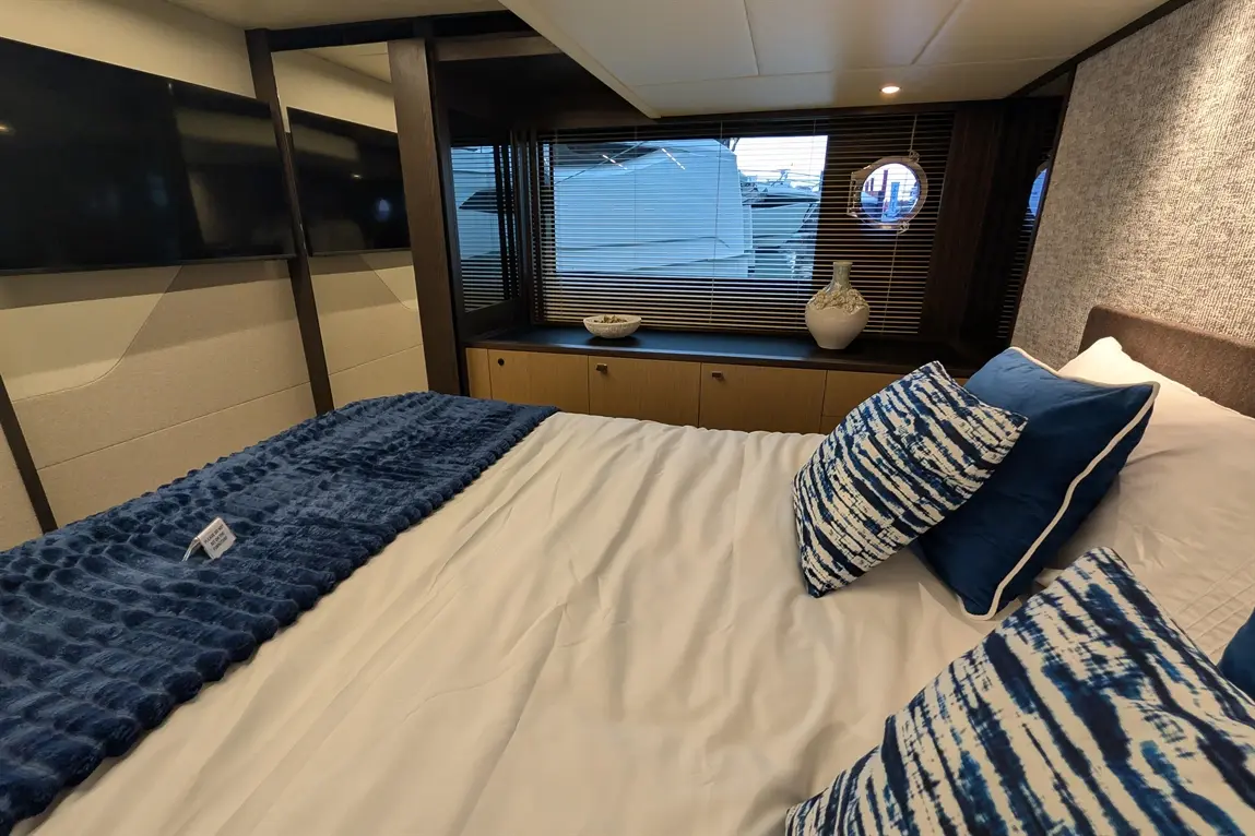Modern bedroom with a blue throw, patterned cushions, two TVs, and a window view of a boat outside.